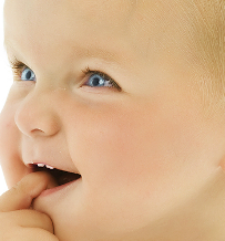 Infant with finger in mouth, looking upward with curious expression against neutral background.