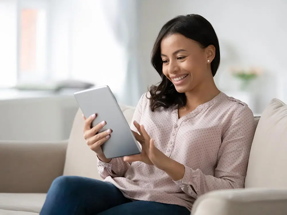 Woman sitting on couch using tablet device in bright living room setting.