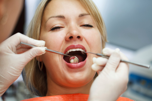 Woman receiving dental examination with mouth open while dentist uses tools in clinical setting.