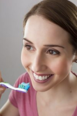 Young woman brushing teeth while smiling at camera, holding pink toothbrush indoors.