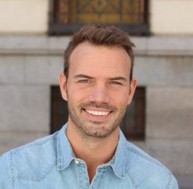 Smiling man in light blue shirt poses for portrait against brick building backdrop.