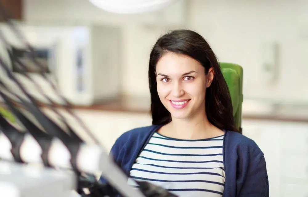 Young woman smiling at camera in modern office setting wearing striped shirt and cardigan.