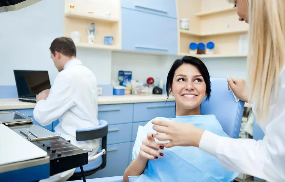 Dental hygienist in blue scrubs smiles while holding tools in modern clinic with colleague working in background.