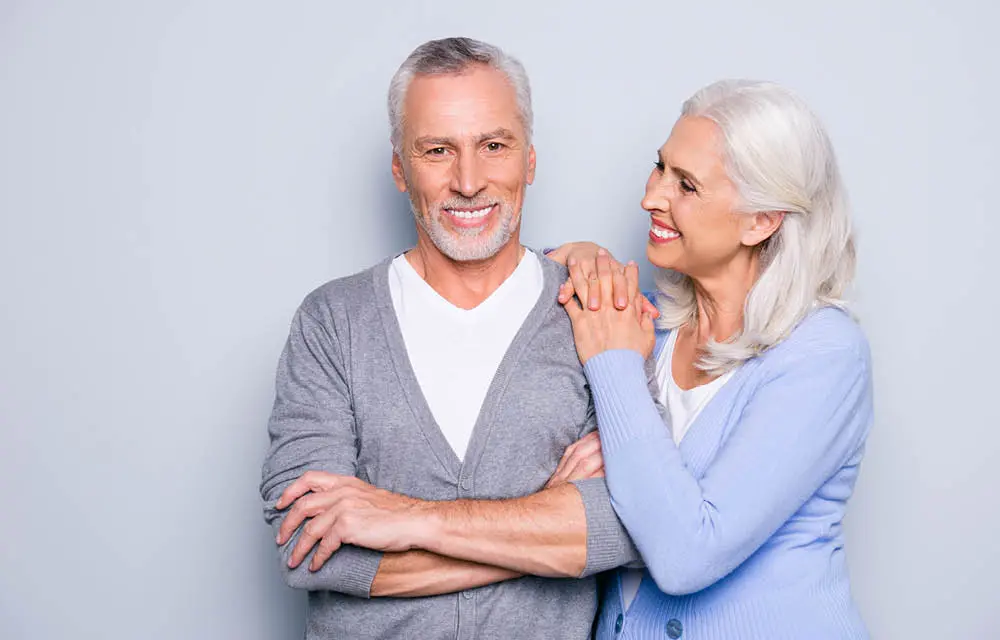 Senior couple smiling together against white background, woman's hand on man's shoulder.