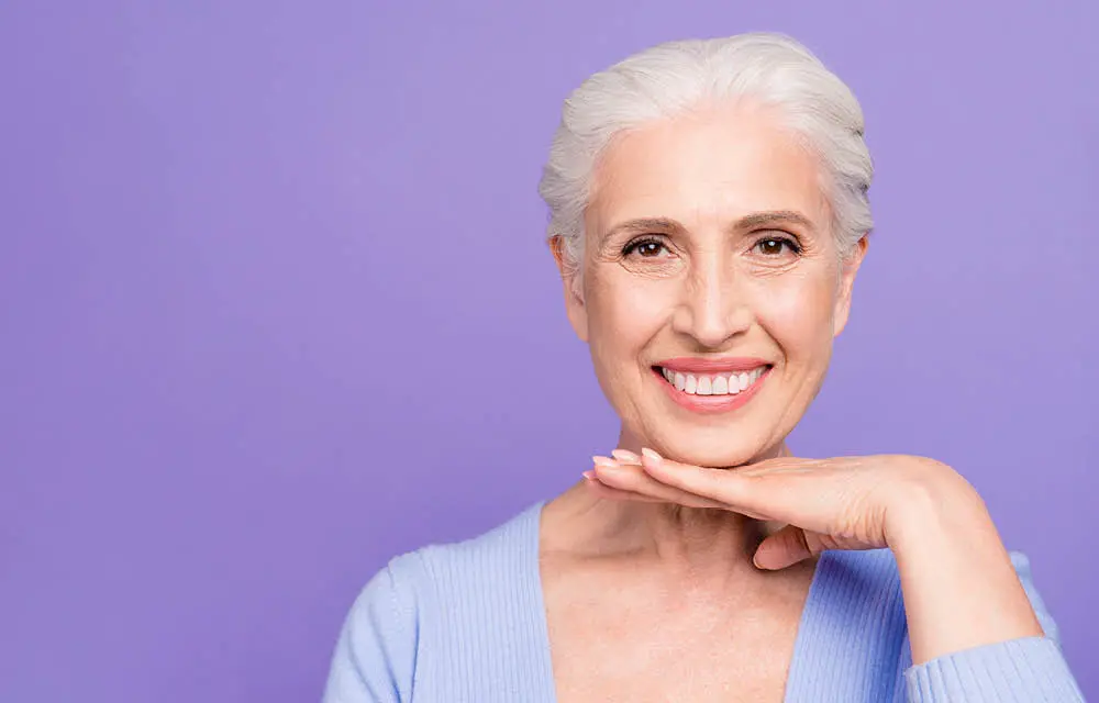 Smiling older woman with white hair against purple background, hand near chin in friendly pose.