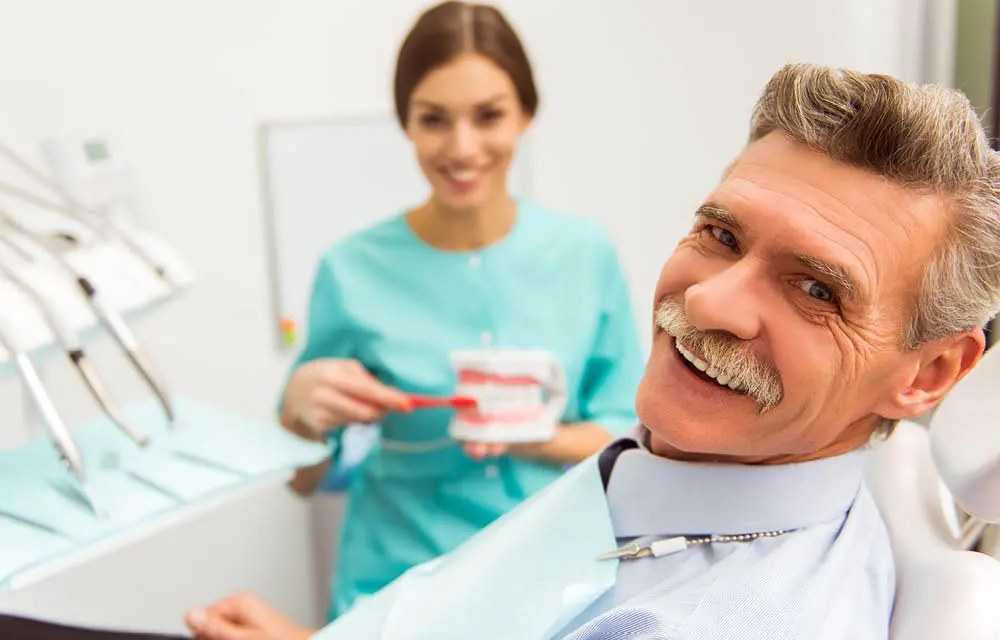 Dentist showing dental model to smiling male patient in modern dental office with assistant.