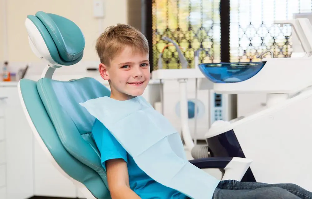 Young boy sitting in blue dental chair holding tablet in modern dental office.