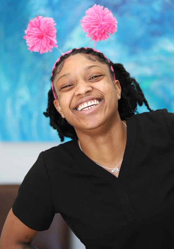 Smiling person wearing black shirt against blue background with pink decorative pom-poms above head.
