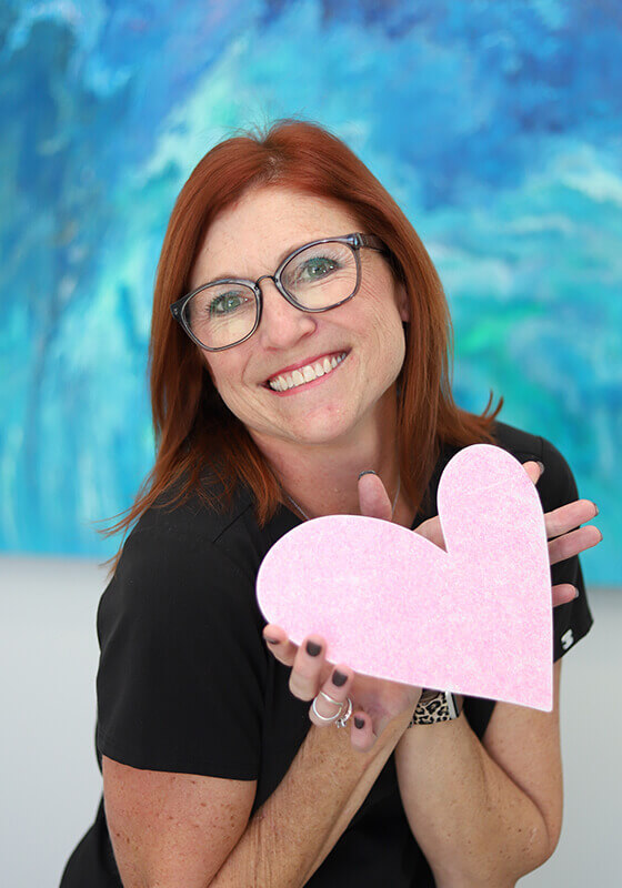 Woman with red hair and glasses holding pink heart shape against blue background.