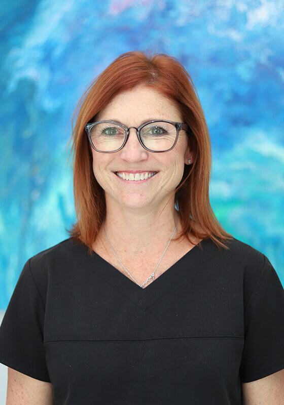 Woman with red hair and glasses smiling at camera against blue abstract background.