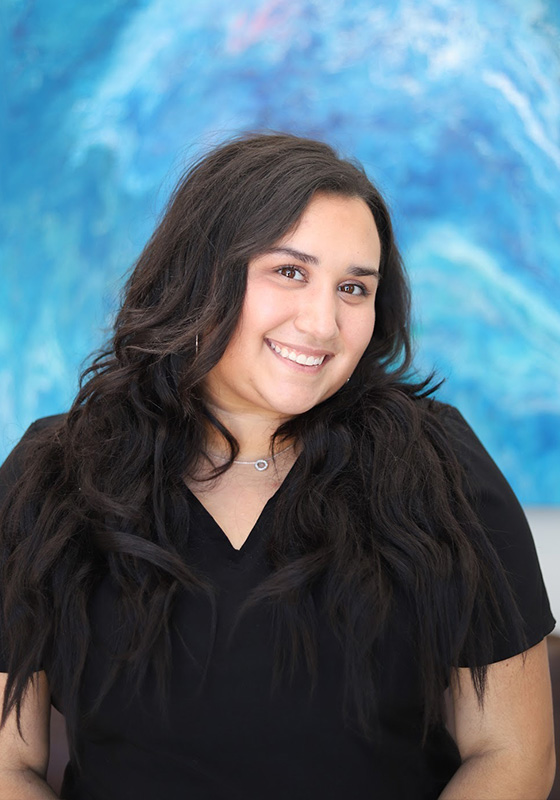 Smiling young woman with long dark hair wearing black shirt against blue background.