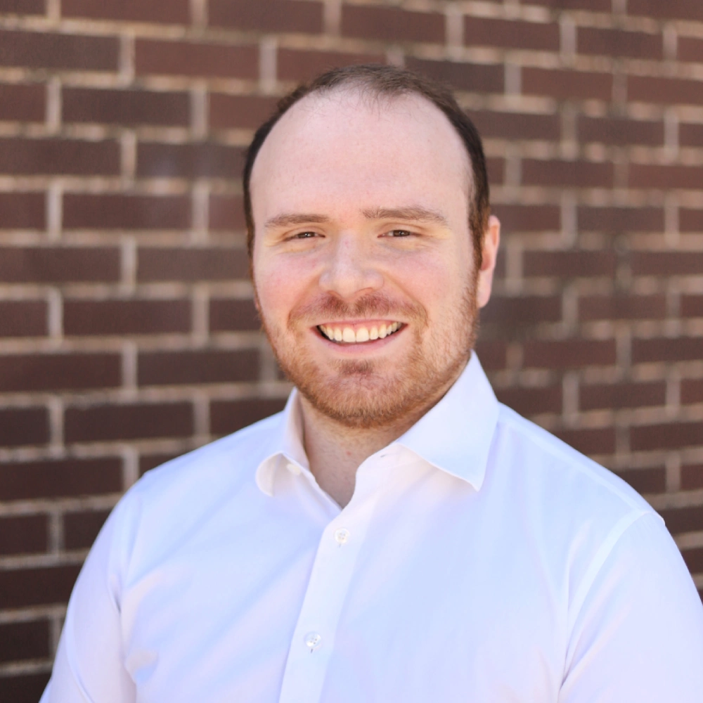 Professional headshot of smiling man in light blue shirt against brick wall background.