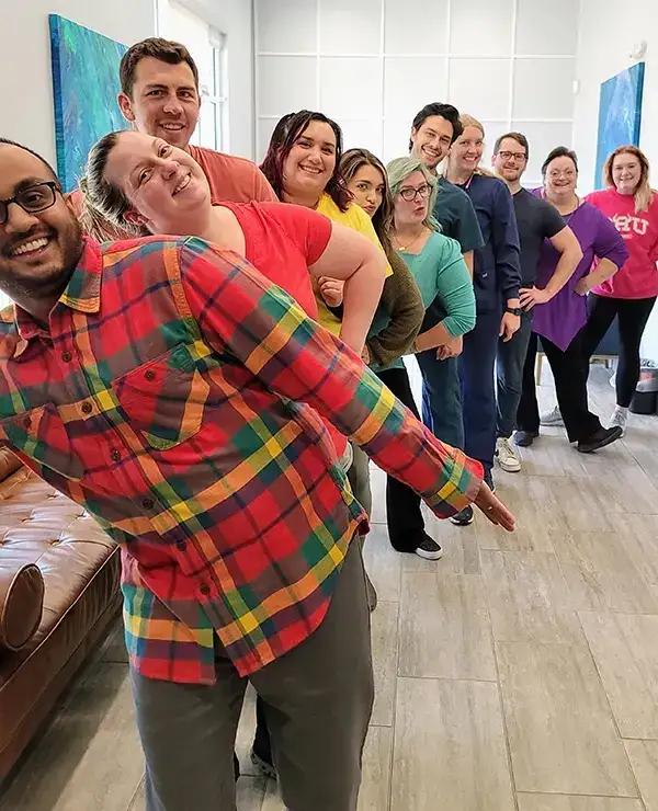 Man in colorful plaid shirt poses playfully while group of people stand together indoors.
