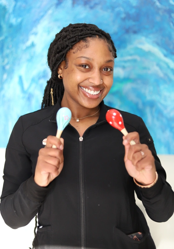 Anaya, Dental Assistant, holding up a light blue maraca and a red polka dot maraca while smiling broadly. She wears a black zip up scrub jacket against a blue textured backdrop.