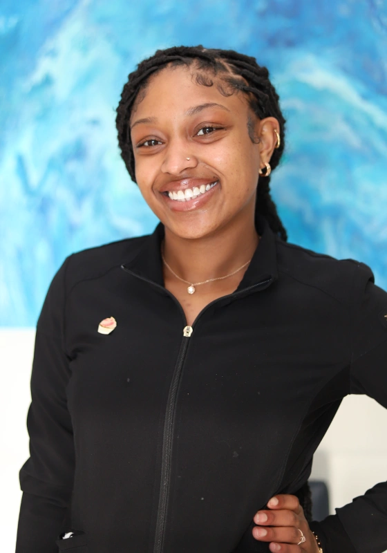 Professional headshot of Anaya, Dental Assistant, smiling with dark locs pulled back, wearing a black scrub jacket with a small pin, standing with one hand on her hip in front of a blue background.