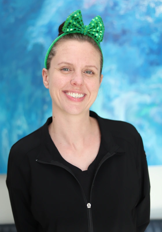 Brittany, Treatment Coordinator, smiling warmly at the camera. She wears a black jacket zip top and a green sequined bow headband against an abstract blue backdrop.