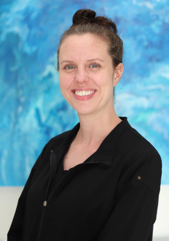 Professional headshot of Brittany, Treatment Coordinator, with her hair styled in a top knot, smiling against an abstract blue wall while wearing a black zip up scrub jacket.