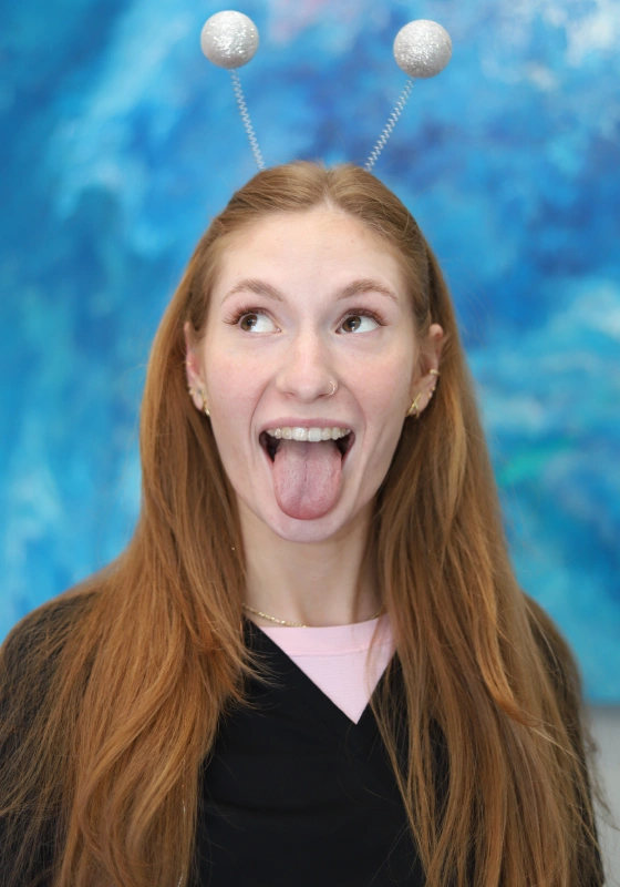 Elaina, Dental Assistant, sticking her tongue out and looking upward while wearing silver glittery alien antennae on a headband. She wears a black top over a light pink undershirt against a blue background.