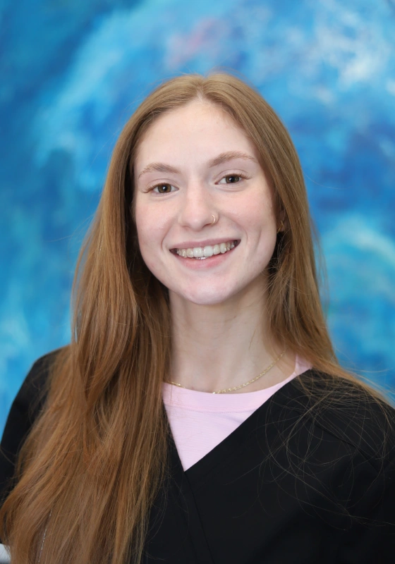 Professional headshot of Elaina, Dental Assistant, with long reddish blonde hair, smiling at the camera in a black scrub top layered over a soft pink tee against an abstract blue backdrop.