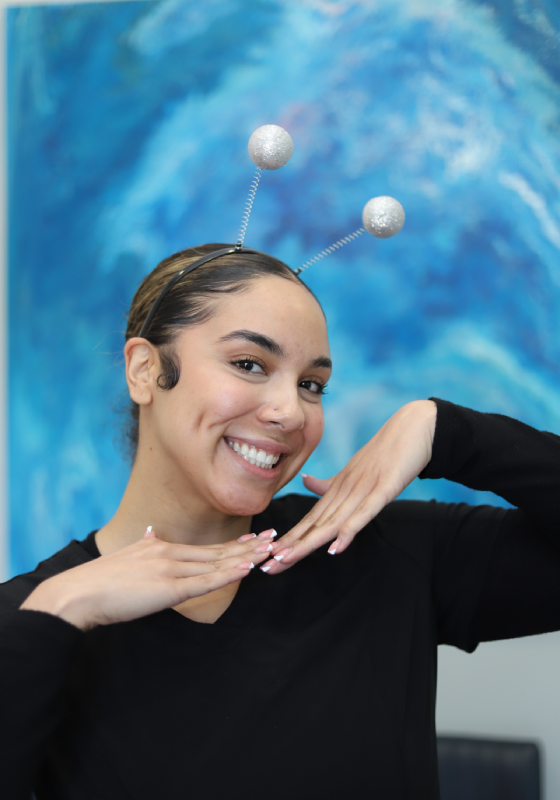 Kyara, Insurance Coordinator, posing playfully with silver glittery alien antennae on a headband. She smiles brightly while framing her chin with both hands, wearing a black top against a vibrant blue abstract backdrop.