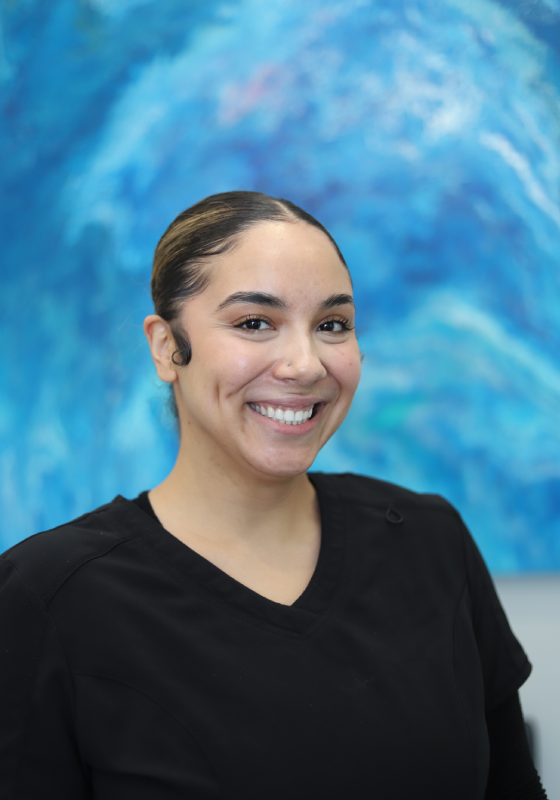 A vertical portrait photo featuring Kyara. She is smiling broadly toward the camera with visible dimples and her hair slicked back into a neat bun with styled edges. She is wearing dark gray or black V neck medical scrubs. The background is an abstract painting composed of swirling motion in vibrant shades of blue, turquoise, and navy.
