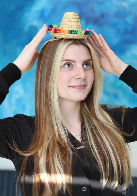 Madie, Dental Assistant, holding a tiny straw sombrero perched on her head with both hands. She has long hair with blonde face framing highlights and wears a black top against a blue textured backdrop.