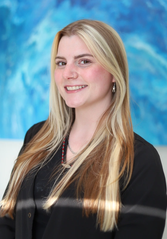 Professional headshot of Madie, Dental Assistant, smiling with her hair down, wearing a black top, a red beaded necklace, and a small silver tooth earring against an abstract blue background.